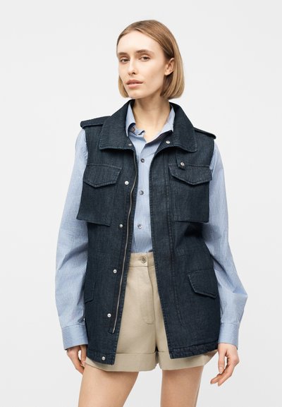 Γυναίκα που φοράει ένα σκούρο τζιν αμάνικο γιλέκο πάνω από ένα μπλε ριγέ πουκάμισο με κουμπιά και μπεζ σορτς με γυρισμένο τελείωμα, στέκεται μπροστά σε ένα απλό φόντο.