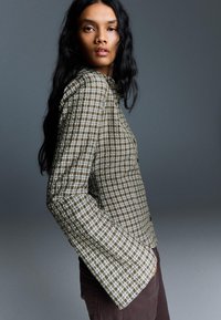 Koszula z długim rękawem, zapinana na guziki, w oliwkowo-zieloną kratę. Charakteryzuje się dopasowanym krojem i szerokimi, rozkloszowanymi rękawami.