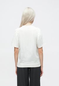 Hvit bomull t-skjorte med puffede ermer og avslappet passform, kombinert med mørkegrå bukser, sett fra bak.