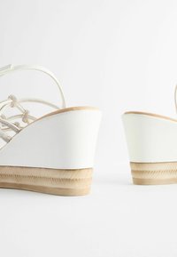Sandales compensées blanches avec une base en bois texturée, présentant un design de brides entrecroisées orné d'accents métalliques.