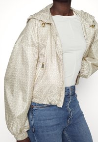 Persona che indossa una giacca a cappuccio beige chiaro a fantasia con zip dorate, una maglietta bianca attillata e jeans blu a vita alta su uno sfondo bianco.
