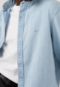 Man in een lichtblauwe Levi's spijkerblouse over een wit T-shirt, met een zilveren ketting en een zwarte riem.