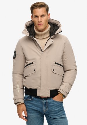Lichtbeige pufferjack met een capuchon, zwarte accenten, twee voorzakken en ribgebreide manchetten. Voorzien van een logo op de mouw en een ritsluiting.