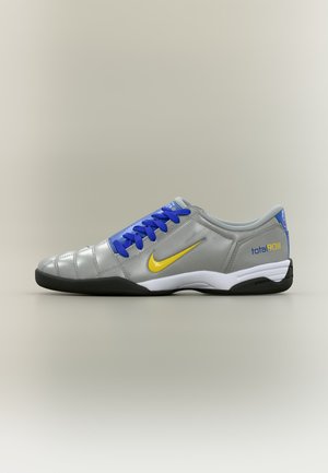 Grauer Nike Total 90 Hallenfußballschuh mit gelbem Swoosh, blauen Schnürsenkeln und schwarzer Sohle auf neutralem Hintergrund.