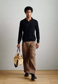 Zwarte, lange mouwen polo; tan kleurige wijde broek; draagt een ronde gele sporttas met bruine accenten; staat op een houten vloer.