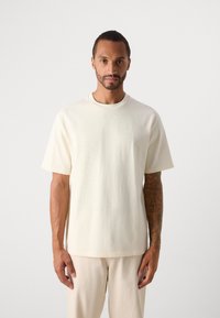 Uomo con capelli corti e tatuaggi in piedi rivolto in avanti, indossando una maglietta oversize bianco sporco e pantaloni abbinati su uno sfondo neutro.