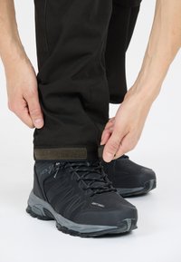 Zwarte wandelschoenen met grijze zolen, bovenwerk van textuurstof en veters. Donkere broek met verstelbare manchetten, vastgezet met een klittenbandsluiting.
