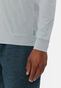 Maglietta grigia chiara a maniche lunghe con scollatura tonda e logo "Vaude" ricamato, abbinata a pantaloni scuri con texture. Presenta un'etichetta verde.