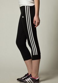 Leggings neri a coste con tre strisce bianche sul lato, vita elastica e design aderente. Indossati con scarpe da ginnastica nere.