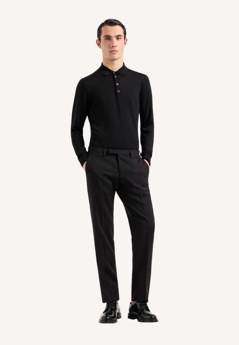 Polo a maniche lunghe nera con tre bottoni, abbinata a pantaloni neri su misura e scarpe nere, in piedi su uno sfondo neutro.