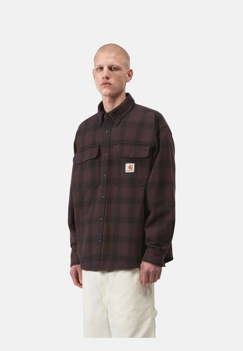 Giovane uomo che indossa una camicia a quadri marrone scuro con bottoni e tasche frontali, e pantaloni chiari, in piedi davanti a uno sfondo semplice.