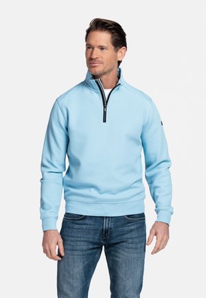 Man in lichtblauwe trui met rits tot kwart hoogte en blauwe spijkerbroek, staand met ontspannen armen, kijkt iets naar de zijkant tegen een witte achtergrond.