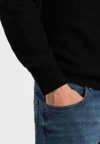 Maglione nero lavorato a maglia con polsini a coste, mostrato con la mano in tasca di jeans blu. La texture è in rilievo e tessuta, creando un interesse visivo.