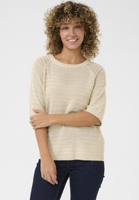 Pull en tricot beige à manches courtes et encolure large, présentant un motif texturé et une coupe décontractée, associé à un jean foncé.
