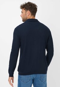 Marineblauwe polo met lange mouwen van een gestructureerde stof en een rechte zoom; gedragen met lichtblauwe jeans die details van het achterdesign tonen.