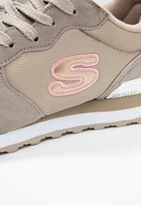 Sneaker beige realizzato in materiali scamosciati e in rete. Presenta una lettera "S" rosa ricamata e una suola intermedia bianca con una suola esterna in gomma testurizzata.