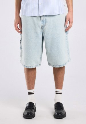 Homme portant un short en jean bleu clair, des chaussettes blanches avec des rayures noires et des mocassins noirs, debout devant un fond clair uni.