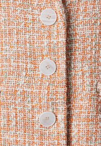 Tvídová látka s kombinací oranžových, bílých a světle zelených nití. Součástí je bílý knoflík se čtyřmi dírkami, přišitý na oděv.