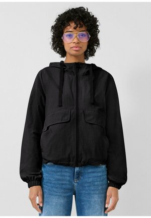 Frau mit lockigem Haar, die übergroße getönte Brillengläser, eine schwarze Kapuzenjacke mit Reißverschluss und blaue Jeans trägt und nach vorne schaut.