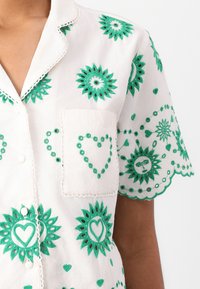 Witte shirt met groene borduursels. Ontwerp bevat bloempatronen, harten en geschulpte mouwen. Detail van de voorzak is zichtbaar.