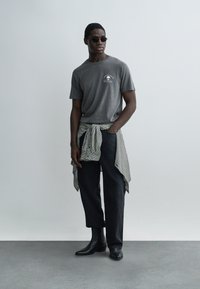 Άνδρας με σκοτεινά γυαλιά ηλίου, γκρι μπλουζάκι με τύπωμα κρανίου, μαύρο παντελόνι, μποτάκια μέχρι τον αστράγαλο, με πουκάμισο καρό δεμένο γύρω από τη μέση του.