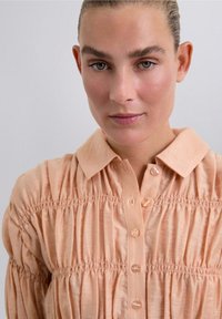 Camisa de color melocotón con cuello tipo polo, cierre de botones y textura recogida a lo largo del torso. Tejido suave con un brillo sutil.