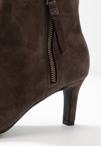 Gros plan d’une botte cheville en daim marron avec une fermeture éclair latérale et un talon noir fin de hauteur moyenne sur une surface blanche.