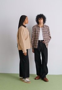 To modeller står side om side. Den ene har på seg en beige oversized jakke med svart krage og høytlivede svarte bukser. Den andre har på seg en brun rutete skjorte over en hvit t-skjorte, med svarte bukser og brune sko.