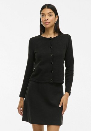 Cardigan nero a coste con scollatura rotonda, maniche lunghe e chiusura con bottoni. Indossato con una gonna nera abbinata, che mette in mostra una silhouette snella.