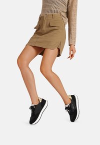 Gonna di denim khaki con tasche frontali e spacco laterale, abbinata a sneakers nere con dettaglio lucido sul tallone e chiusura con lacci.