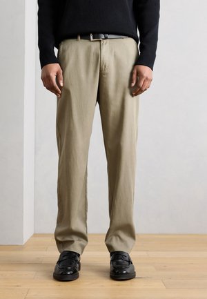 Homme portant un pantalon beige, une ceinture noire, un haut à manches longues noir et des chaussures en cuir noires, debout sur un sol en bois.