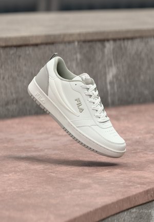 Witte en grijze FILA sneaker met veters, zwevend boven een roze betonnen oppervlak, met een grijze stenen muur op de achtergrond.