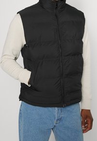 Gilet isolante nero con colletto alto, zip frontale e due tasche laterali, indossato sopra una camicia a maniche lunghe di colore chiaro.