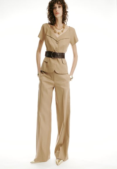 Woman wearing a beige short-sleeve blazer with large buttons, wide-legged pants, a wide dark belt, gold necklace, bracelet, and pointed gold shoes.