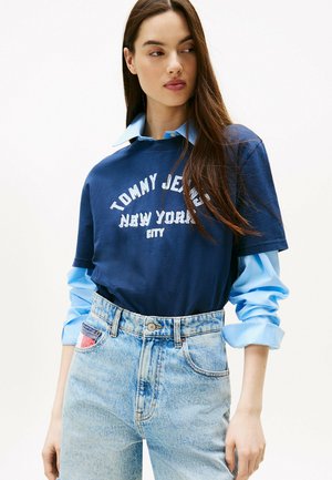 T-shirt oversize bleu marine avec l'imprimé "TOMMY JEANS NEW YORK CITY", superposé sur une chemise boutonnée bleu clair, associé à un jean bleu clair taille haute.