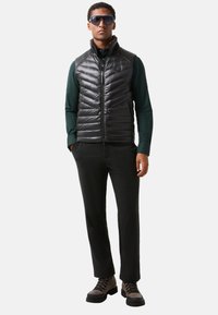 Zwarte puffer vest met een geborduurd ontwerp, gedragen over een donkergroene shirt. Zwarte broek en bruine wandelschoenen, aangevuld met oversized zonnebril.