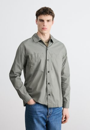 Νέος άνδρας με μαλλιά τύπου μπούκλα φοράει ανοιχτό γκρι πουκάμισο με κουμπιά πάνω από μπεζ εσωτερικό μπλουζάκι και μπλε τζιν, στέκεται μπροστά σε απλό φόντο.