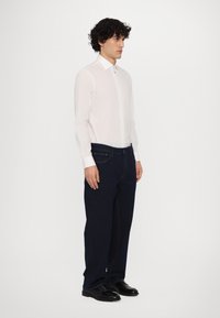 Camisa blanca de manga larga con cuello abotonado, combinada con jeans azul oscuro y zapatos negros. Los jeans tienen bolsillos frontales visibles y costuras.