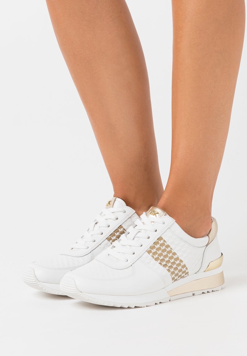 Jambes portant des baskets blanches avec des accents dorés et des panneaux latéraux à motifs, debout sur une surface blanche unie.