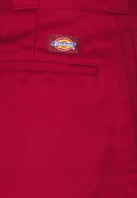 Červená látka s hladkým povrchem, v horní kapse je přišitá hnědá nášivka s logem "Dickies" ve žluté a červené barvě.