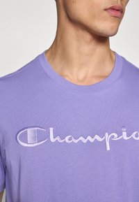 Μωβ βαμβακερό t-shirt με κεντημένο λογότυπο "Champion" σε πιο ανοιχτή απόχρωση, με κλασικό στρογγυλό λαιμό και μαλακή υφή.