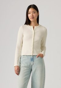 Ριπ ζακέτα σε κρεμ χρώμα με μακριά μανίκια και στρογγυλή λαιμόκοψη, με κουμπιά μπροστά και κοντή εφαρμογή, συνδυασμένη με ξεβαμμένο μπλε τζιν υψηλής μέσης.