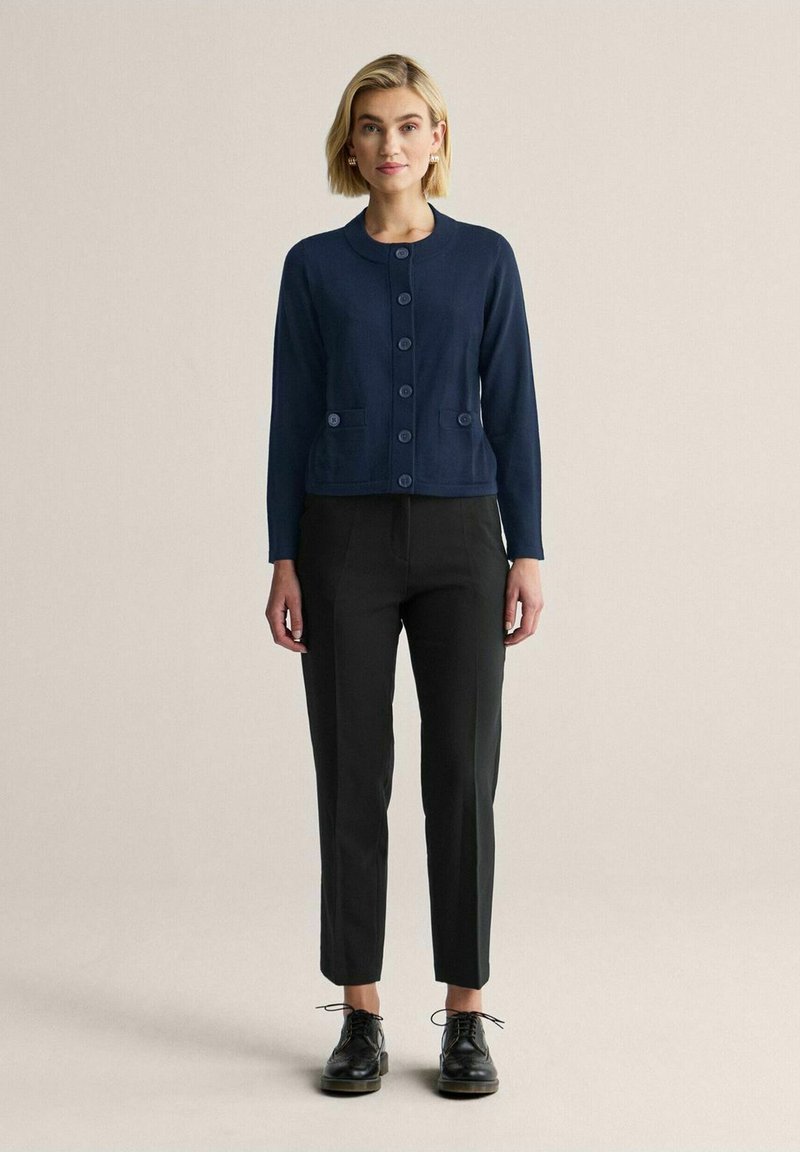 Marinblå stickad kofta med rund hals, knappar fram och två påsydda fickor. Stylist med svart tajta byxor och svarta skor.