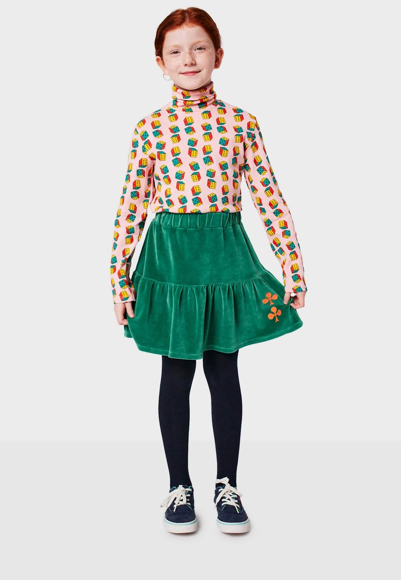 Jupe en velours vert avec un ourlet volanté, associée à un col roulé à manches longues ornée de motifs colorés, ainsi qu'à des collants navy et des baskets.
