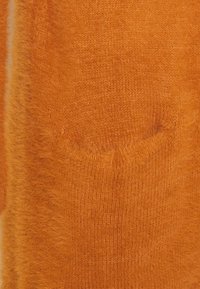 Pomarańczowa dzianina o miękkiej, puszystej fakturze. Zawiera detal kieszeni, ukazujący puszysty wygląd przędzy i żywy kolor.