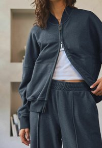 Donkergrijze sportjack met een rits aan de voorkant, geribde manchetten en een losse pasvorm, gecombineerd met bijpassende losse broek en een witte tanktop eronder.