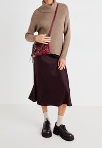 Femme portant un pull col roulé beige, une jupe midi violette foncée, des chaussures volumineuses sombres, des chaussettes crème et un petit sac bandoulière rouge.