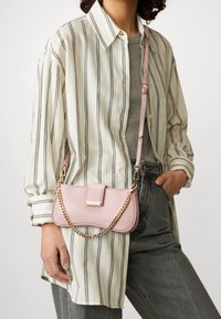 Różowa skórzana torba na ramię ze złotym łańcuszkowym paskiem, o prostokątnym kształcie, z klapką i ozdobnym detalem, zestawiona z koszulą w paski i dżinsami.