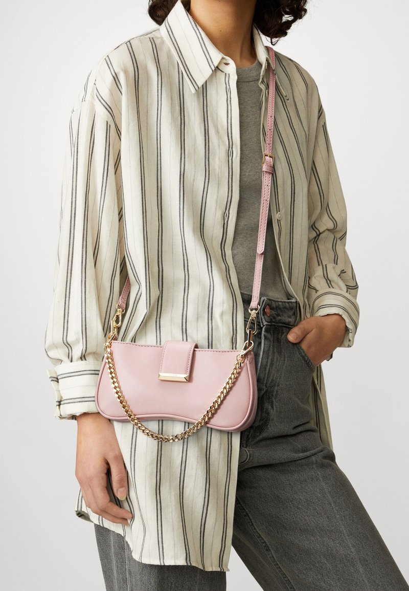 Różowa skórzana torba na ramię ze złotym łańcuszkowym paskiem, o prostokątnym kształcie, z klapką i ozdobnym detalem, zestawiona z koszulą w paski i dżinsami.