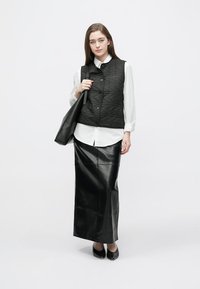 Svart quiltet vest over en hvit skjorte med knapper, kombinert med et langt svart lærskjørt og svarte sko. Modellen holder en svart tote-veske.